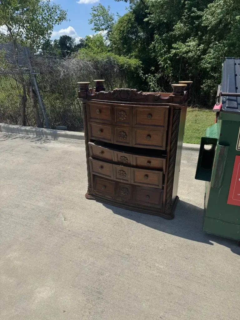 An ornate wooden dresser left on the pavement beside a commercial dumpster