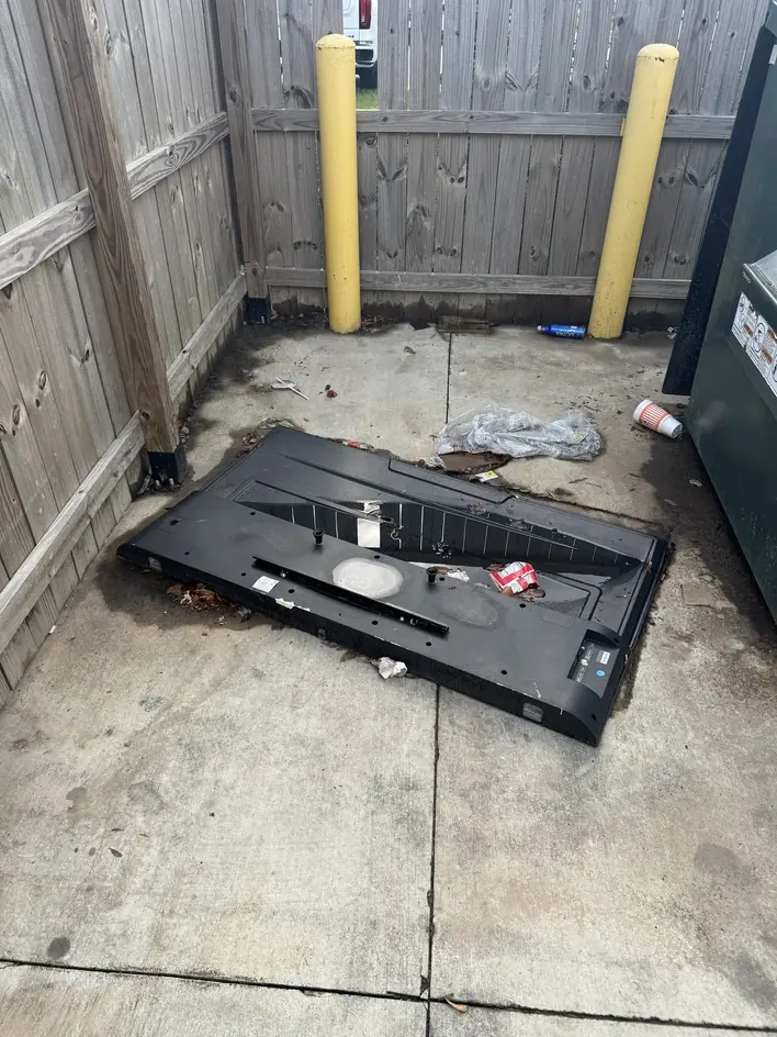 A large flat-screen television broken and abandoned on the concrete of a dumpster enclosure