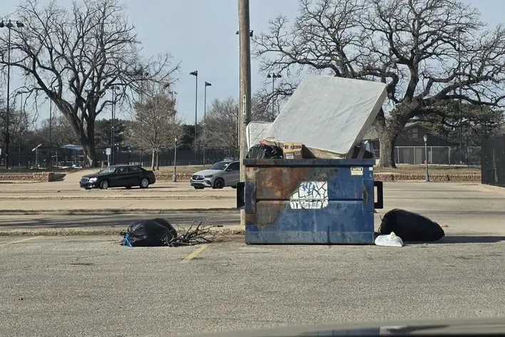 A dumpster in a parking lot with bags and loose debris scattered on the ground around it