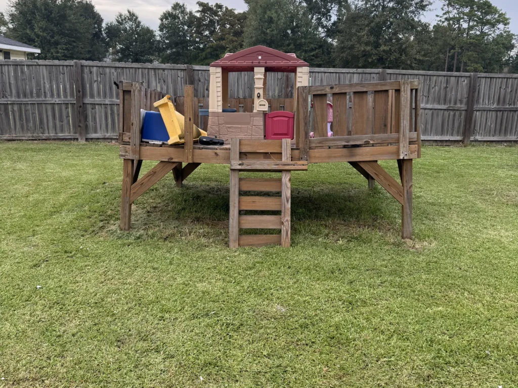 A raised wooden playset in a fenced Moss Bluff back yard before removal